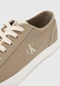 Baskets en toile beige avec semelle en caoutchouc blanche, lacets crème et un logo "CK" subtil de couleur claire sur le côté. Texture lisse.