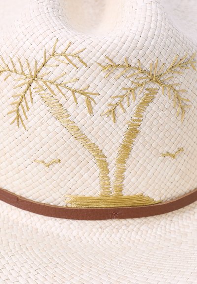 Woven straw hat in cream with palm tree embroidery in gold, featuring a brown leather band around the base. Textured surface with patterned details.