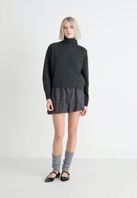 Suéter de cuello alto de punto verde oscuro con bajo acanalado, combinado con una falda a rayas gris y negra. Modelo lleva calentadores de piernas grises y bailarinas negras.