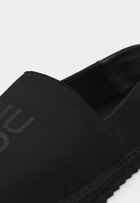 Chaussure noire à enfiler avec semelle texturée et logo embossé sur le dessus, montrant les détails latéraux et supérieurs sur un fond uni.