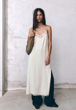 Frau mit langen dunklen Haaren, die ein cremefarbenes Unterkleid über weite dunkle Jeans trägt, hält eine olivgrüne Tasche mit Nieten und steht vor einer strukturierten Wand.