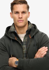 Felpa verde scuro con zip, dotata di un colletto a quadri. La felpa presenta una texture liscia e dettagli in pelle. È visibile un orologio da polso.