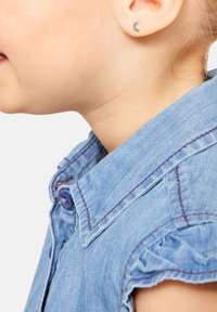 Camicia di denim di colore azzurro chiaro, con maniche a volant e cuciture viola. Piccole e colorate orecchini a forma di mezzaluna adornano l'orecchio.