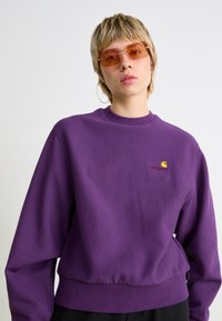 Lilla sweatshirt med ribbet halsudskæring og manchetter, med et lille broderet logo på brystet. Glat tekstur, afslappet pasform.