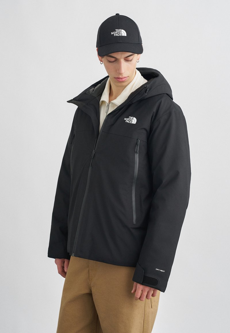 Zwarte waterdichte jas met een capuchon, ritssluitingen en het North Face-logo op de borst. Draag het met beige broek en een zwarte pet.