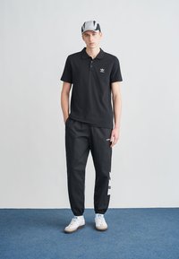 Černé polo tričko s bílým logem, doplněné černými tepláky s bočními pruhy. Outfit doplňují bílé tenisky.