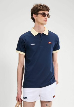 Jeune homme portant un polo bleu marine avec col beige et short blanc, tenant un sac beige, lunettes de soleil foncées, debout devant un fond uni.