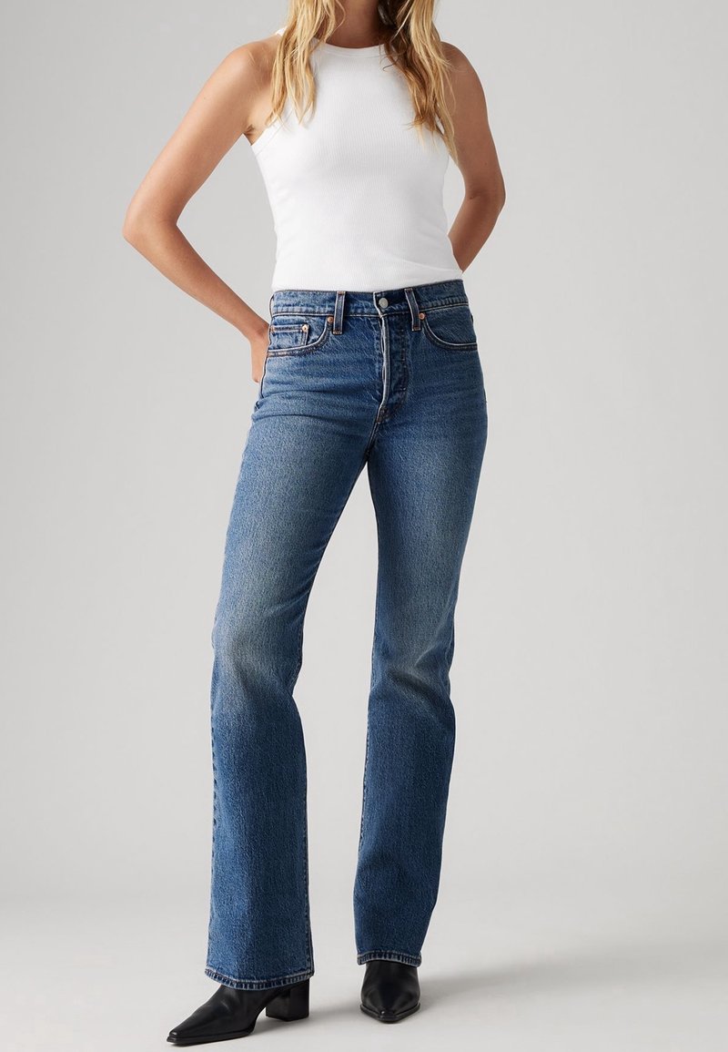 Vaqueros acampanados de mezclilla azul con cintura alta, acabado desgastado y diseño de cinco bolsillos, combinados con una camiseta blanca sin mangas y botas negras de tacón.