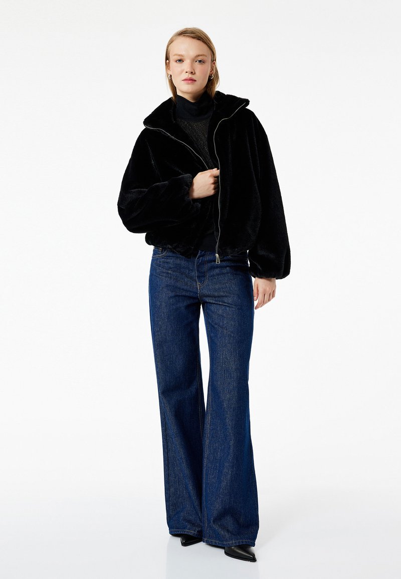 Chaqueta de piel sintética negra con cremallera y cuello oversize, llevada sobre un suéter oscuro y combinada con unos jeans de mezclilla azul de tiro alto y pierna acampanada.