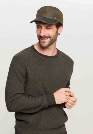 Mann, der lächelt und einen strukturierten, dunkeloliven Pullover sowie eine gesteppte, olivfarbene Kappe mit einem dunkelgrauen Schirm trägt, mit locker verschränkten Händen.