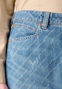 Närbild av en person som bär ljusblå slitna jeans och en beige långärmad skjorta, med fokus på jeansfickan och sömmarna.