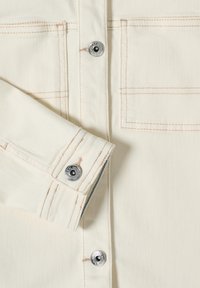 Gros plan sur une veste en denim crème avec poches poitrine, coutures contrastantes et boutons-pression métalliques sur le poignet et la patte de boutonnage avant.