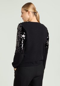 Maglione nero con maniche con paillettes, collo rotondo, corpo strutturato e vestibilità comoda. Indossato con pantaloni neri.
