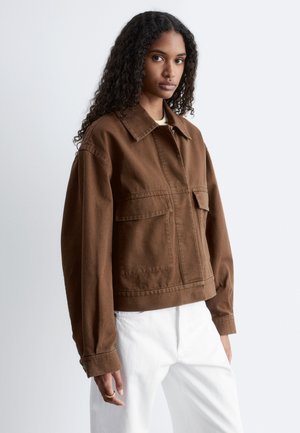 Jonge vrouw met lang krullend haar, een bruine jas met knopen en witte broek draagend, staand tegen een effen grijze achtergrond.