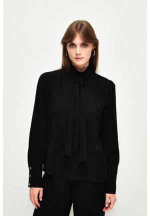Black blouse with long sleeves, a tied collar, pleated details, and gold-button accents on the cuffs. Smooth fabric texture.