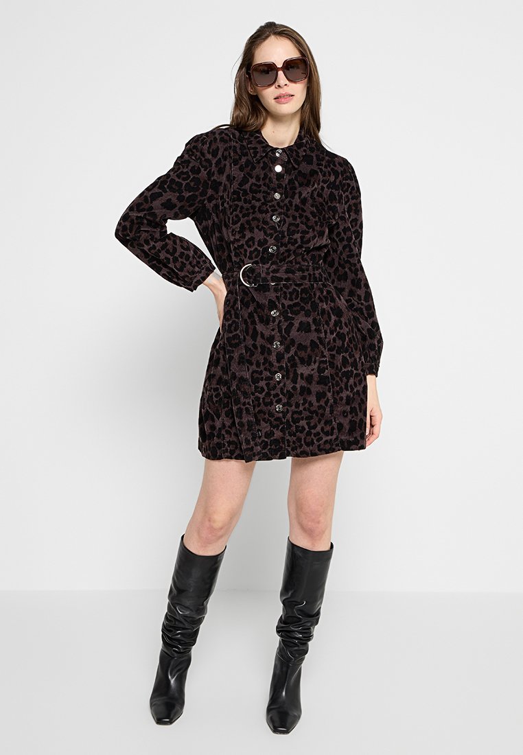 Donna che indossa un vestito a stampa leopardata scura con cintura, stivali in pelle nera fino al ginocchio e occhiali da sole oversize, in piedi con una mano sulla vita.
