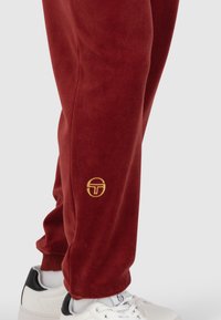 Rotvelours-Jogginghosen mit einem goldenen, gestickten Logo am Oberschenkel, kombiniert mit weißen Sneakern mit schwarzen Akzenten.