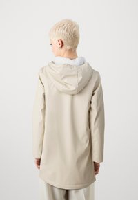 Lys beige regnfrakk med hette, med myk hvit fleecefôr, lange ermer og forlenget bakhem for dekning.
