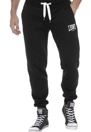 Modèle portant un pantalon de survêtement noir Leone avec des cordons blancs et le logo, associé à des baskets montantes noires.