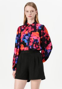 Blomsterbluse med livlige rosa, blå og oransje mønstre, med krage og puffete ermer. Kombinert med høyt liv svarte shorts.