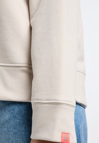 Beige sweatshirt met een gladde textuur, voorzien van een geribbelde manchet en een klein roze merklabel aan de onderkant, gecombineerd met blauwe jeans.
