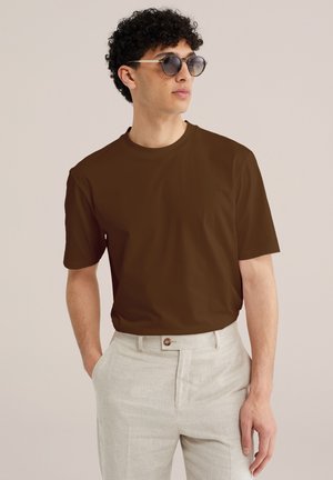 Mann mit runder Sonnenbrille, braunem Kurzarmhemd und hellbeiger Hose, stehend mit einer Hand in der Tasche vor einfarbigem Hintergrund.