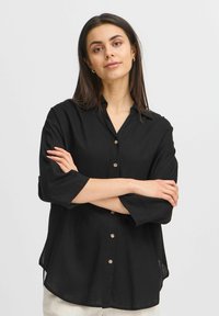 Camicia nera button-up realizzata in tessuto leggero, con colletto, maniche lunghe e orlo curvo. Piccole bottoni marroni decorano la parte anteriore.