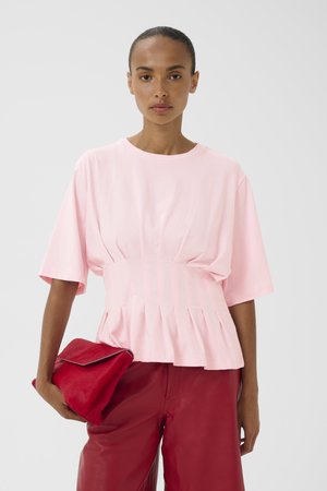 Kvinde iført en lyserød plisseret kortærmet top og røde læderbukser, der holder en rød clutchtaske foran en neutral grå baggrund.