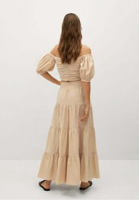 Beige off-shoulder mekko, jossa on puffi hihnat, rypytetty vartalo ja kerroksellinen hame. Sileä rakenne, lattiaa hipova muotoilu ja minimaalinen yksityiskohtaisuus.