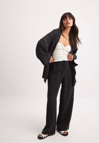 Zwarte, loszittende broek gecombineerd met een zwarte kimono-achtige jas over een witte, geribbelde strapless top; model draagt sandalen met bandjes.