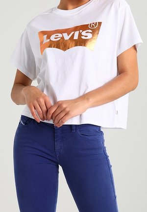 Person wearing a white Levi's crop top with a metallic logo and high-waisted blue jeans, hands loosely clasped in front.