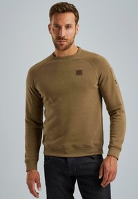 Olivgrüner Sweatshirt aus weichem Stoff, mit Rundhalsausschnitt, Raglanärmeln und einem kleinen Aufnäher auf der Brust. Dunkle Jeans runden den Look ab.