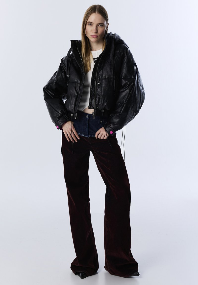 Vrouw in een zwarte cropped pufferjack, witte crop top, donkerrode fluwelen wijde broek en zwarte puntige schoenen voor een effen achtergrond.