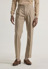 Pantalones beige a medida con frente plano, cierre de dos botones y dobladillos con puños, hechos de una tela suave, combinados con zapatos de ante marrón.