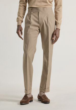 Pantalones beige a medida con frente plano, cierre de dos botones y dobladillos con puños, hechos de una tela suave, combinados con zapatos de ante marrón.