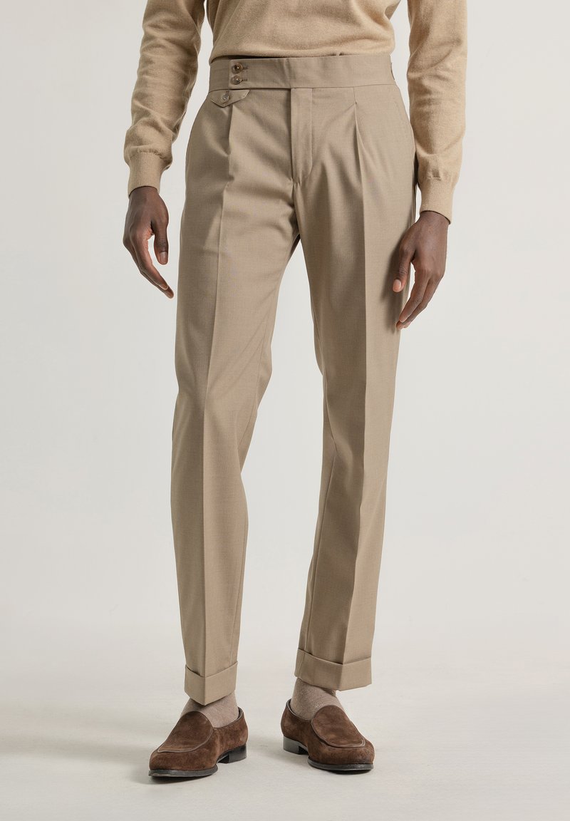 Pantalones beige a medida con frente plano, cierre de dos botones y dobladillos con puños, hechos de una tela suave, combinados con zapatos de ante marrón.