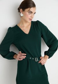 Tmavozelené šaty s výstrihom do V a balónovými rukávmi. Obsahujú opasok s striebornými prvkami a hladkú textúru látky.