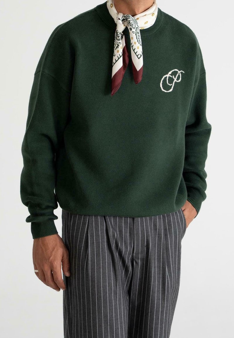 Homme portant un pull vert foncé avec un logo brodé blanc, une écharpe blanche à motif autour du cou, et un pantalon gris à fines rayures avec la main dans la poche.