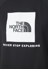 Logo The North Face v černé barvě na bílém čtverci s frází "Nikdy nepřestávejte prozkoumávat" níže na černém textilním pozadí.