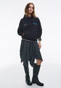 Fekete kapucnis pulóver türkiz hímzéssel, barna felsőréteg fölött, szürke aszimmetrikus szoknyával és térdig érő fekete csizmával párosítva.