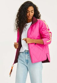 Prošívaná růžová bunda se žlutými bočními pruhy, zipem vpředu a hnědou podšívkou. Obsahuje dekorativní táhlo zipu a dvě boční kapsy.