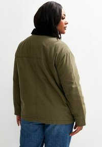 Olivgrön jacka med svart krage, med avslappnad passform, synliga sömmar och en slät textur, sedd framifrån.