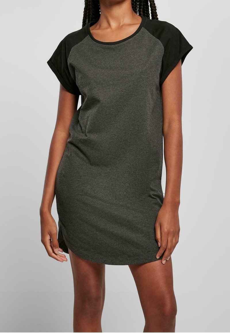 Femme portant une robe gris foncé à manches courtes avec des manches raglan noires, debout devant un fond clair uni.