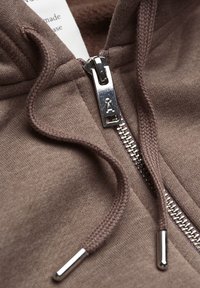 Brun huvtröja med metalldragkedja och dragsko, närbild som visar dragkedjans dragkrok och en del av tyglabeln inuti.