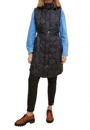 Vrouw draagt een lange zwarte gewatteerde mouwloze bodywarmer over een blauwe blouse, zwarte broek en bruine chunky veterlaarzen.