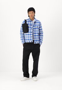 Blauw-wit geruite jas met een zwarte pouch, gecombineerd met zwarte broek en witte sneakers. Glad materiaal, comfortabele pasvorm en knoopdetails.