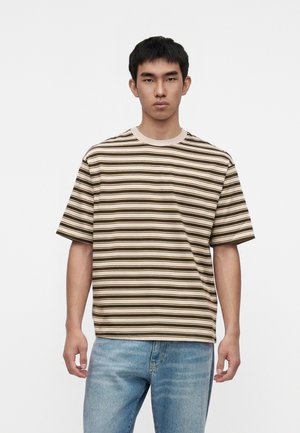 Homme portant un t-shirt beige et marron à rayures horizontales et un jean bleu clair, debout devant un fond blanc.