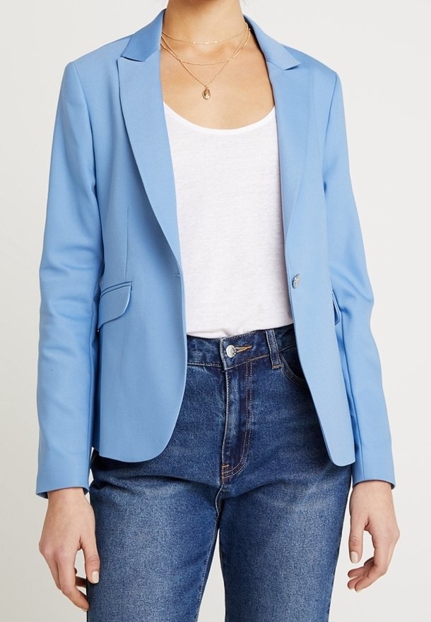 Femme portant un blazer bleu clair sur un débardeur blanc et un jean bleu, avec des colliers dorés superposés.