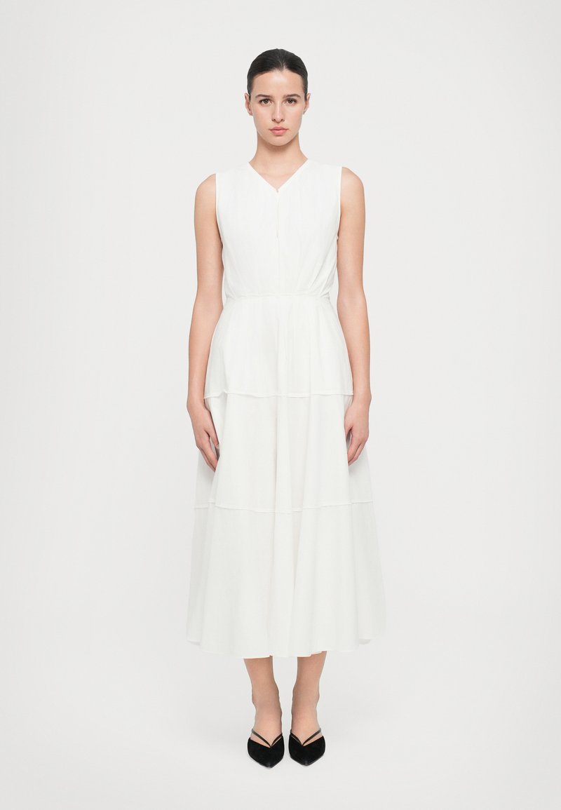 White sleeveless dress with a V-neck, featuring a tiered design and a cinched waist. Smooth fabric and a flowing silhouette. Black shoes complete the look.