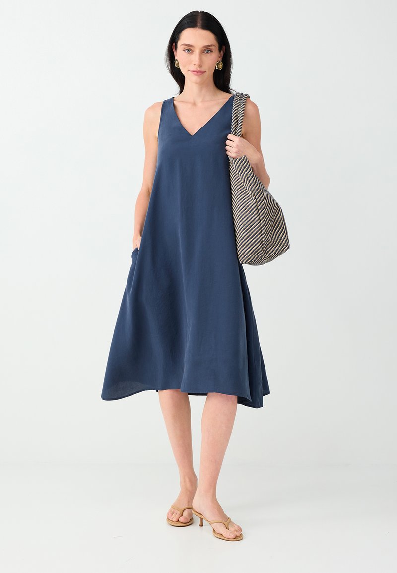 Femme en robe midi bleu marine sans manches, portant un grand sac à bandoulière rayé et des sandales à talons beiges avec des boucles d'oreilles dorées.